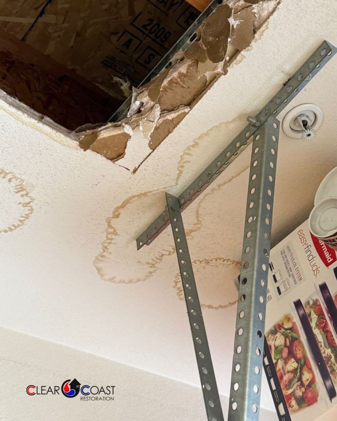 Damaged ceiling with hole, water stains, and metal support brackets. Clear Coast Restoration logo.