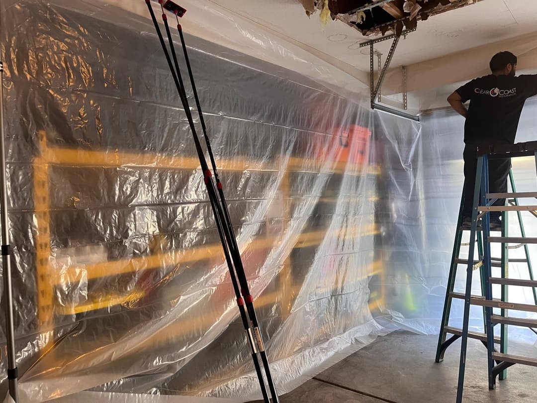 Man working on a ladder behind a plastic barrier in a renovation project, with shelves visible.