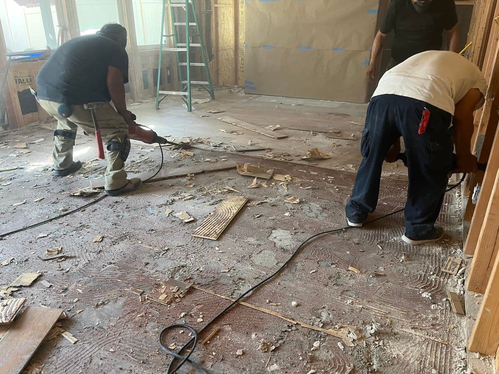 Workers renovating a room, removing flooring and debris, with tools and equipment visible.