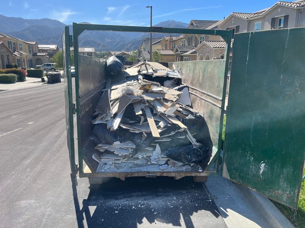 Construction debris in a dumpster on a residential street with mountains in the background.