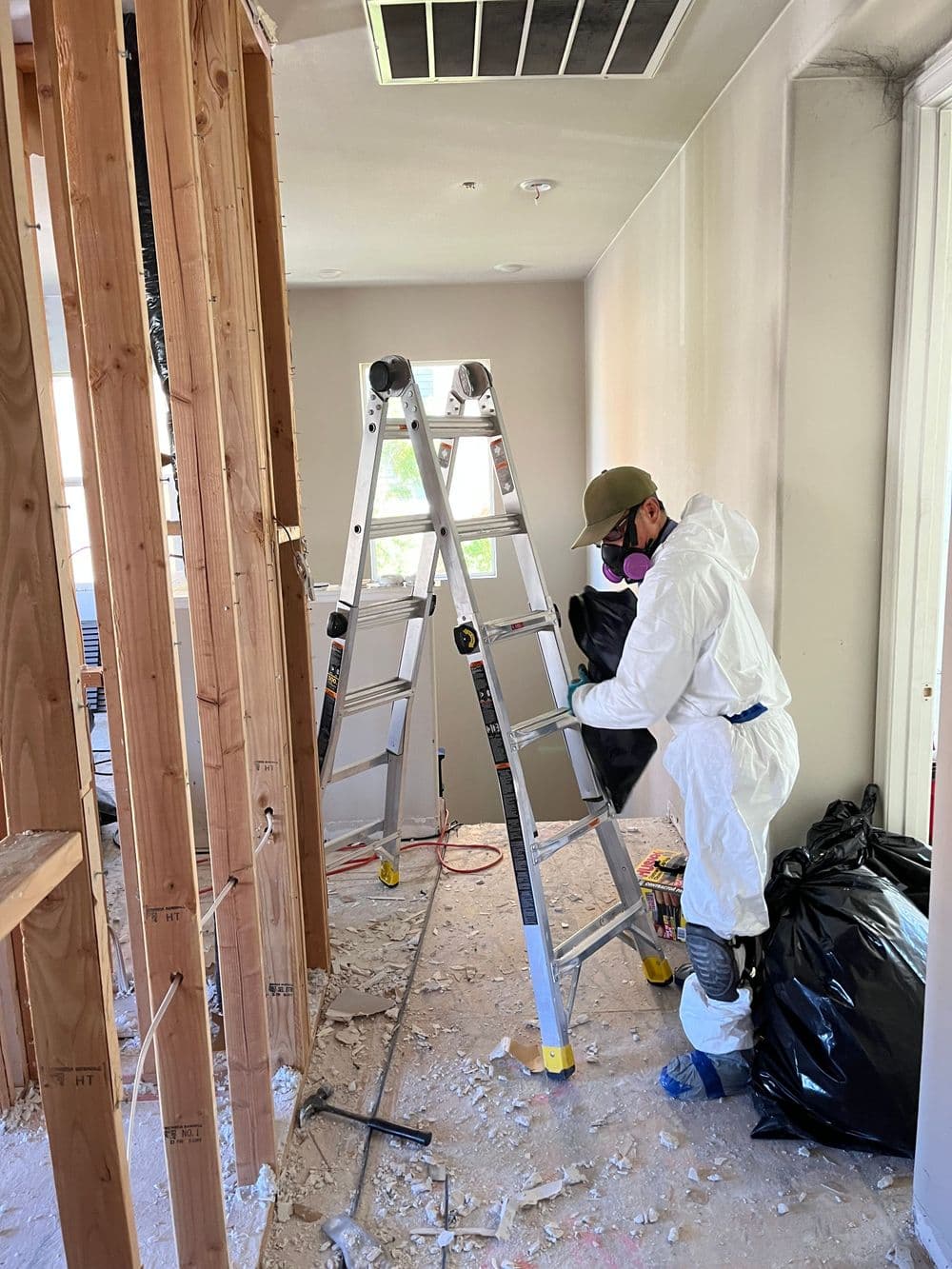 Person in protective gear cleaning debris in a renovated room with a ladder and trash bags.