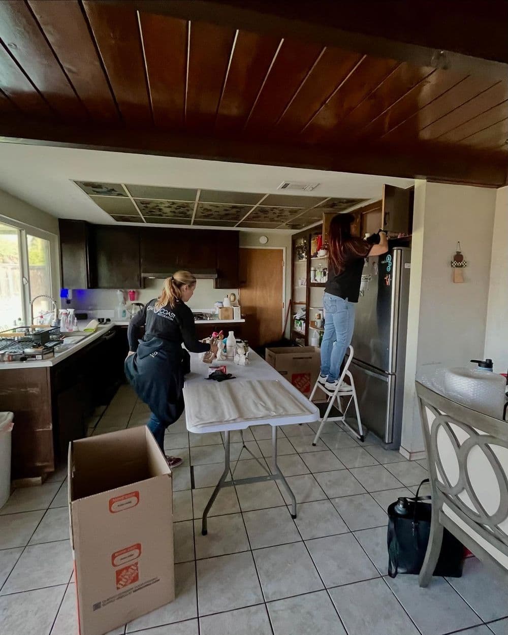 Two women organizing a modern kitchen, one on a ladder and another at a table with supplies.
